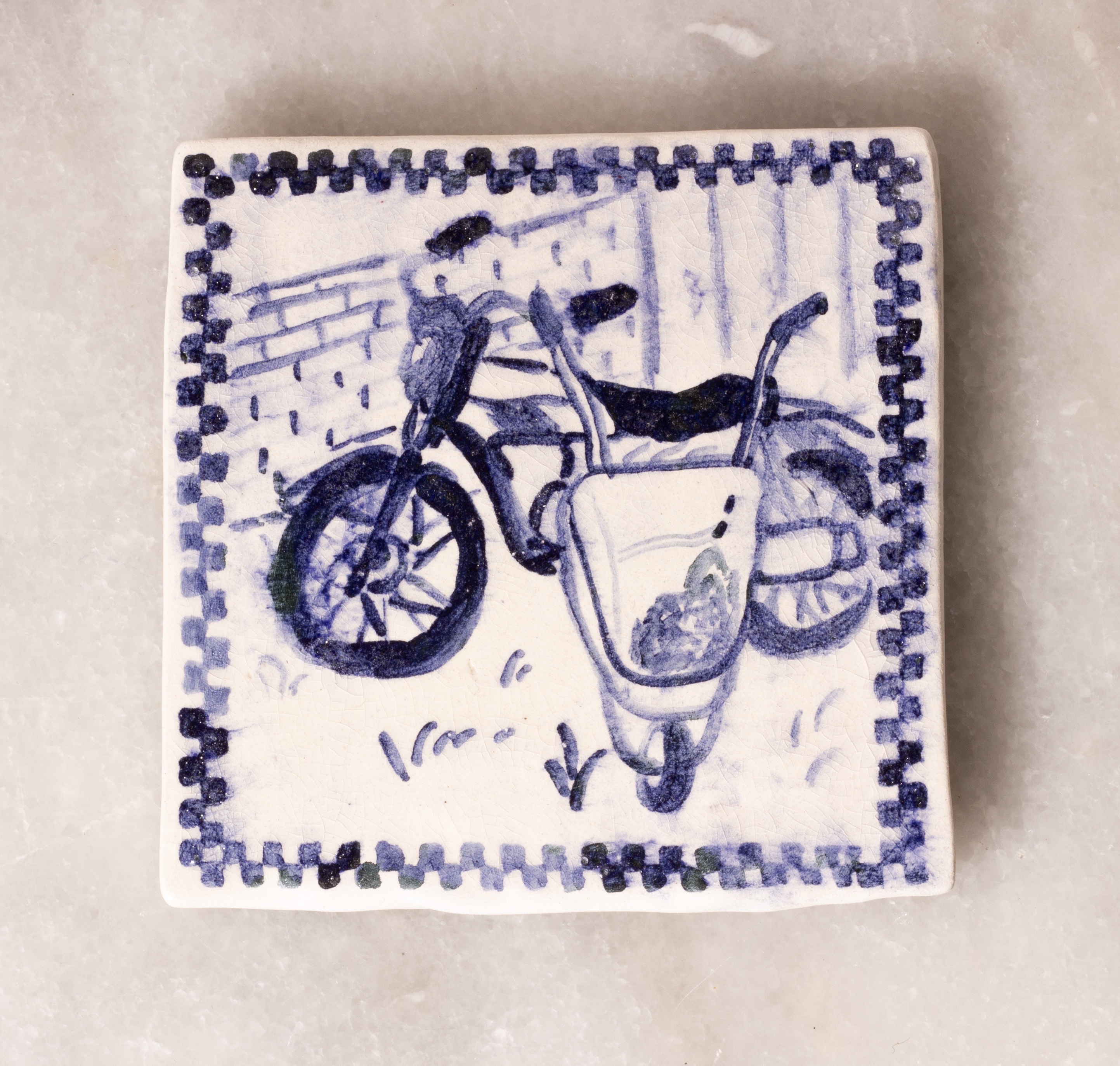 carreau de faïence blanc peint en bleu à l'oxyde de cobalt. Il représente une brouette, posée devant une mobylette, avec un mur en pierre en fond