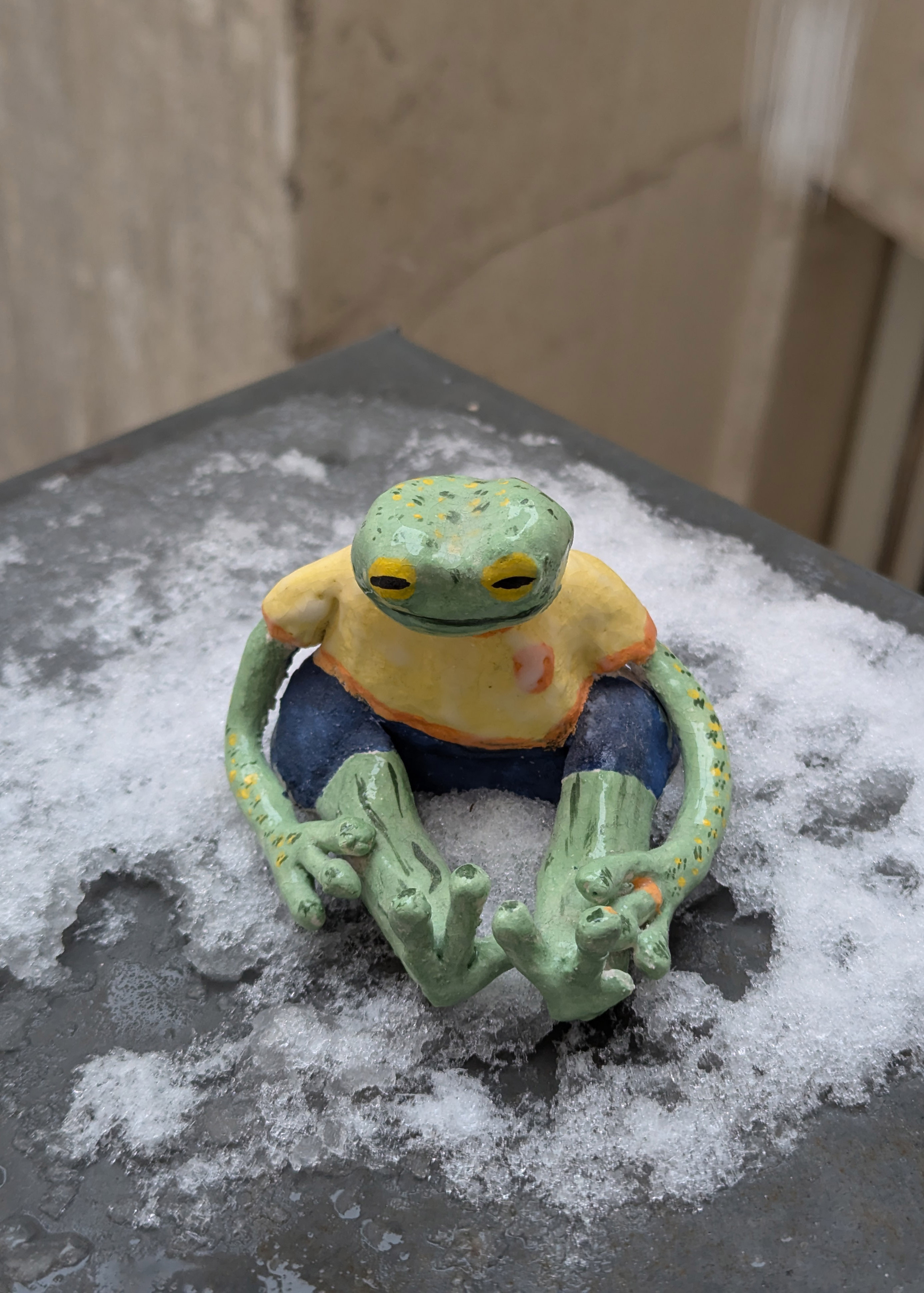grenouille verte à tâches jaunes en faïence assise, elle porte un t-shirt jaune à col orange et un jean, elle sourit